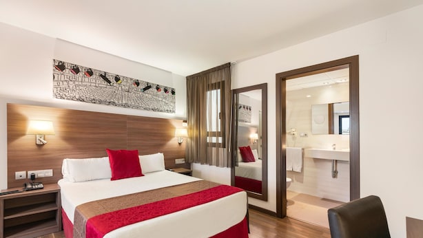 A hotel room with a large bed, a wooden headboard, a red throw blanket, a nightstand with a lamp, a mirror, and a bathroom visible through an open door. - Hotel Best Auto Hogar (Barcelona)