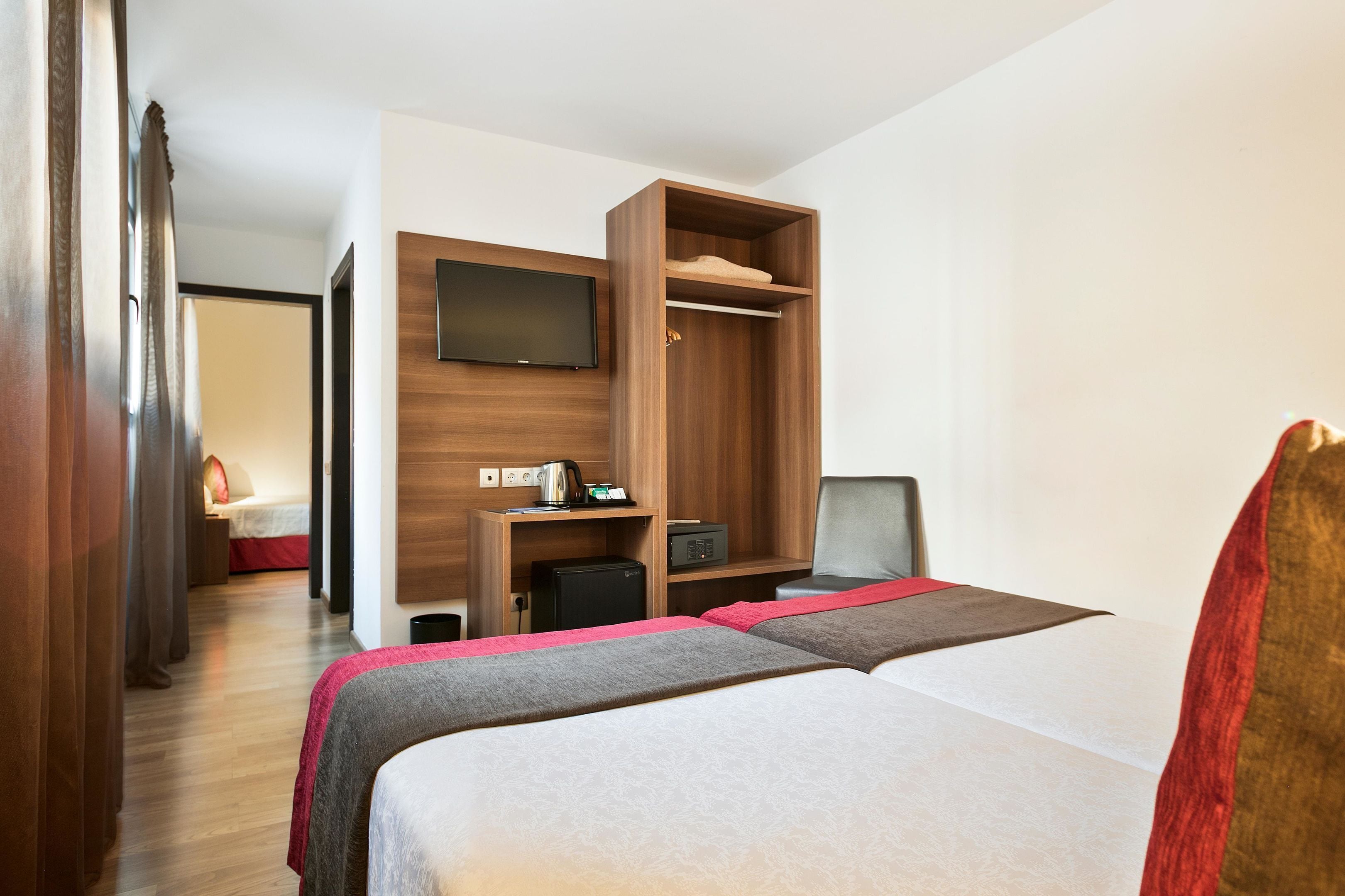 A hotel room with two beds, a TV mounted on the wall, a desk with a computer, and a wardrobe.