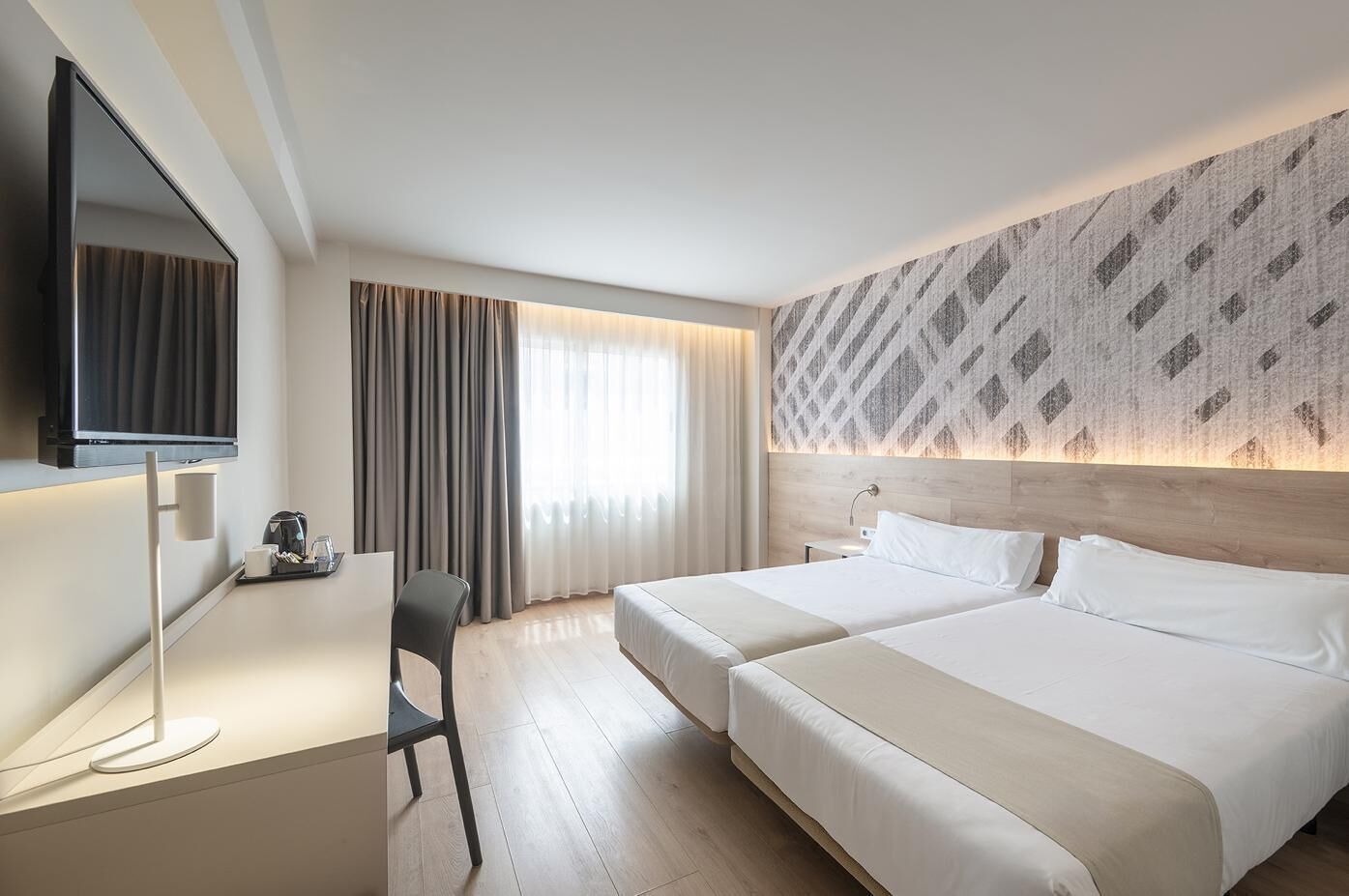Habitación de hotel con dos camas, un escritorio, un televisor y un ventanal con cortinas.