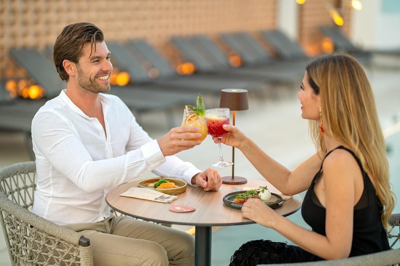 Two people seated at a round table outdoors, holding drinks.