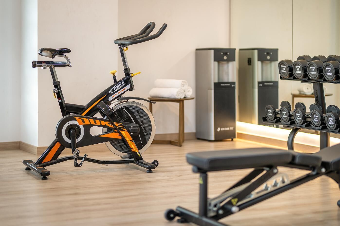 A stationary bike, a bench, and a rack of dumbbells in a room with a wooden floor and a mirror.