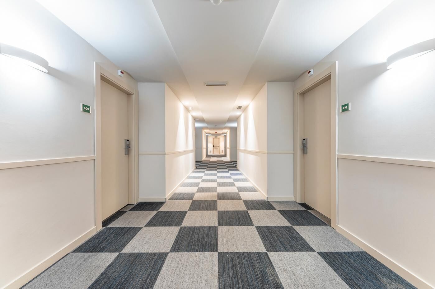 Een hotelgang met een geruit tapijt, lichte wanden en deuren voorzien van kamernummers.