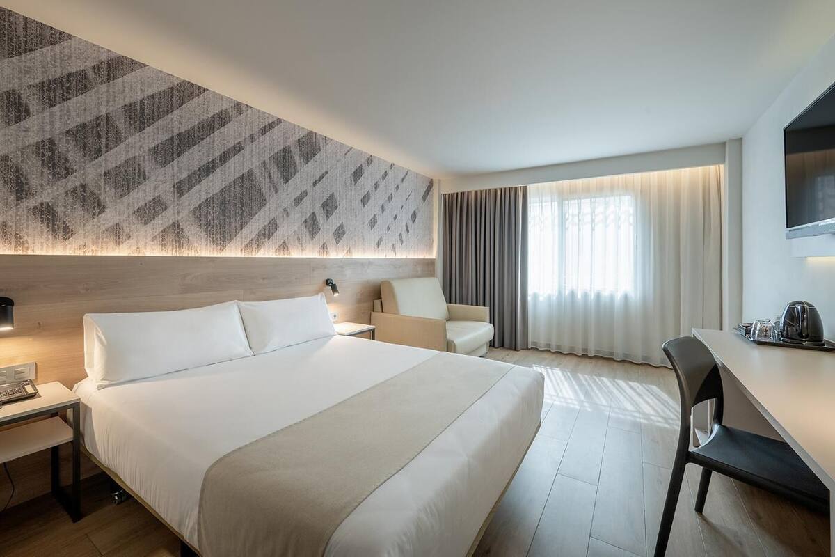 Una habitación de hotel moderna con una cama grande, un escritorio, una silla, un televisor y un ventanal con cortinas.