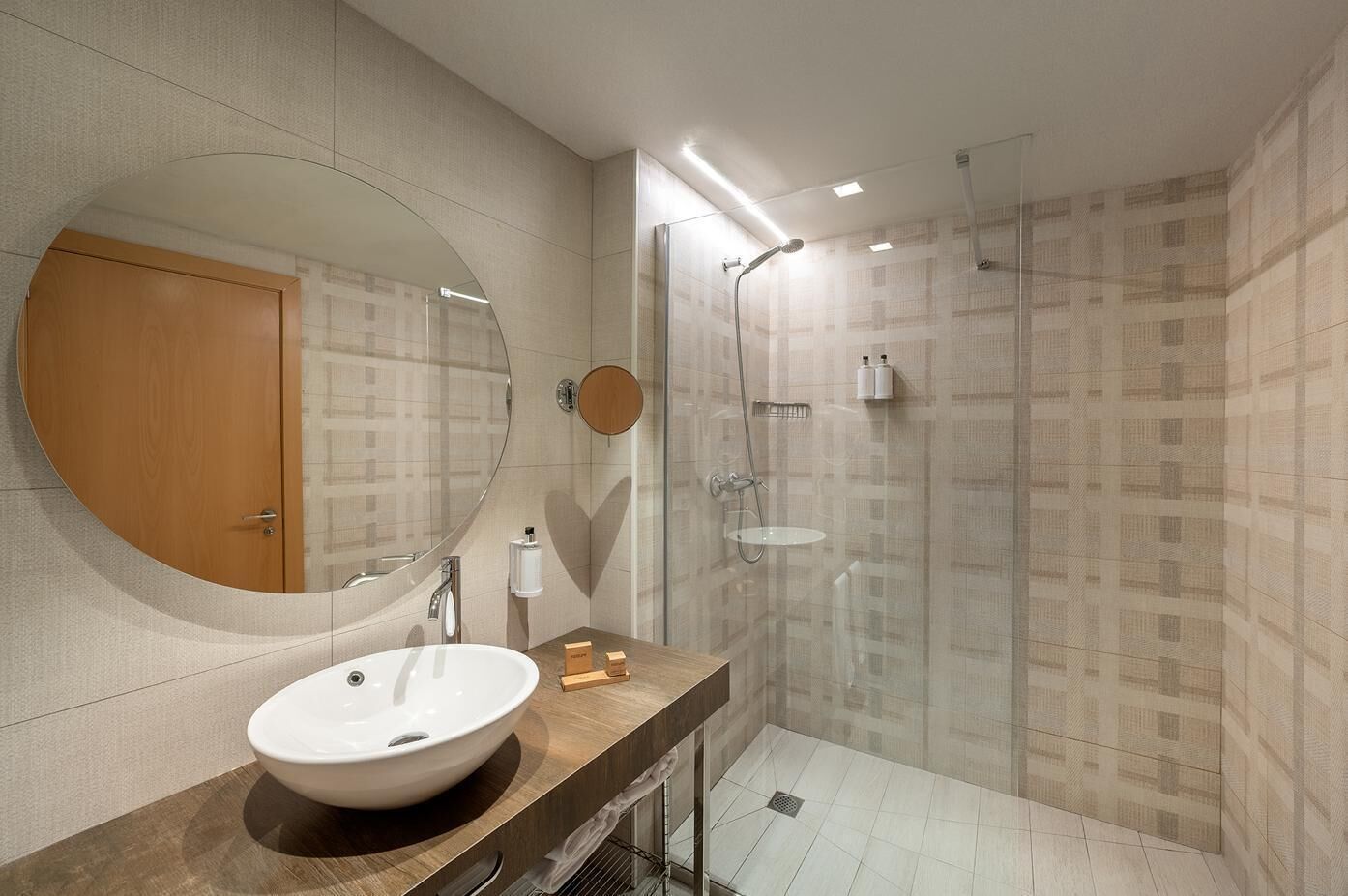 A modern bathroom with a large oval mirror, a wooden vanity, a white sink, a glass-enclosed shower, and patterned walls.