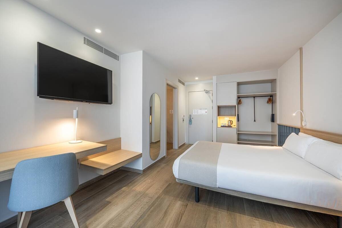 Una habitación de hotel moderna con cama, escritorio con silla, televisor, espejo y armario.