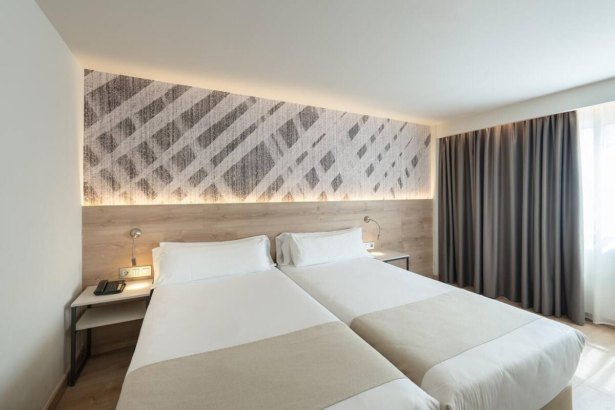 Habitación de hotel con dos camas, cabecera de madera, pared con estampado, mesita de noche con lámpara y ventana con cortinas.