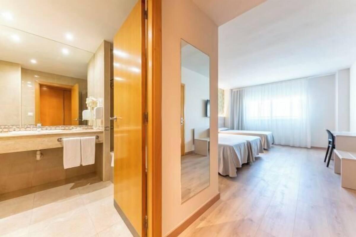 Una habitación de hotel con un espejo grande, una puerta de madera, un baño con tocador y una cama con colcha blanca.