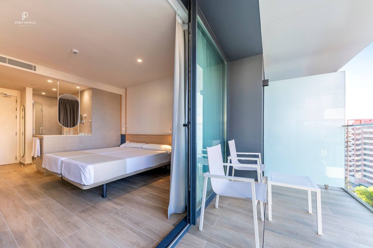 Una habitación de hotel moderna con una cama, una mesita y sillas. Hay una puerta corrediza de vidrio que da a un balcón con vistas a edificios.