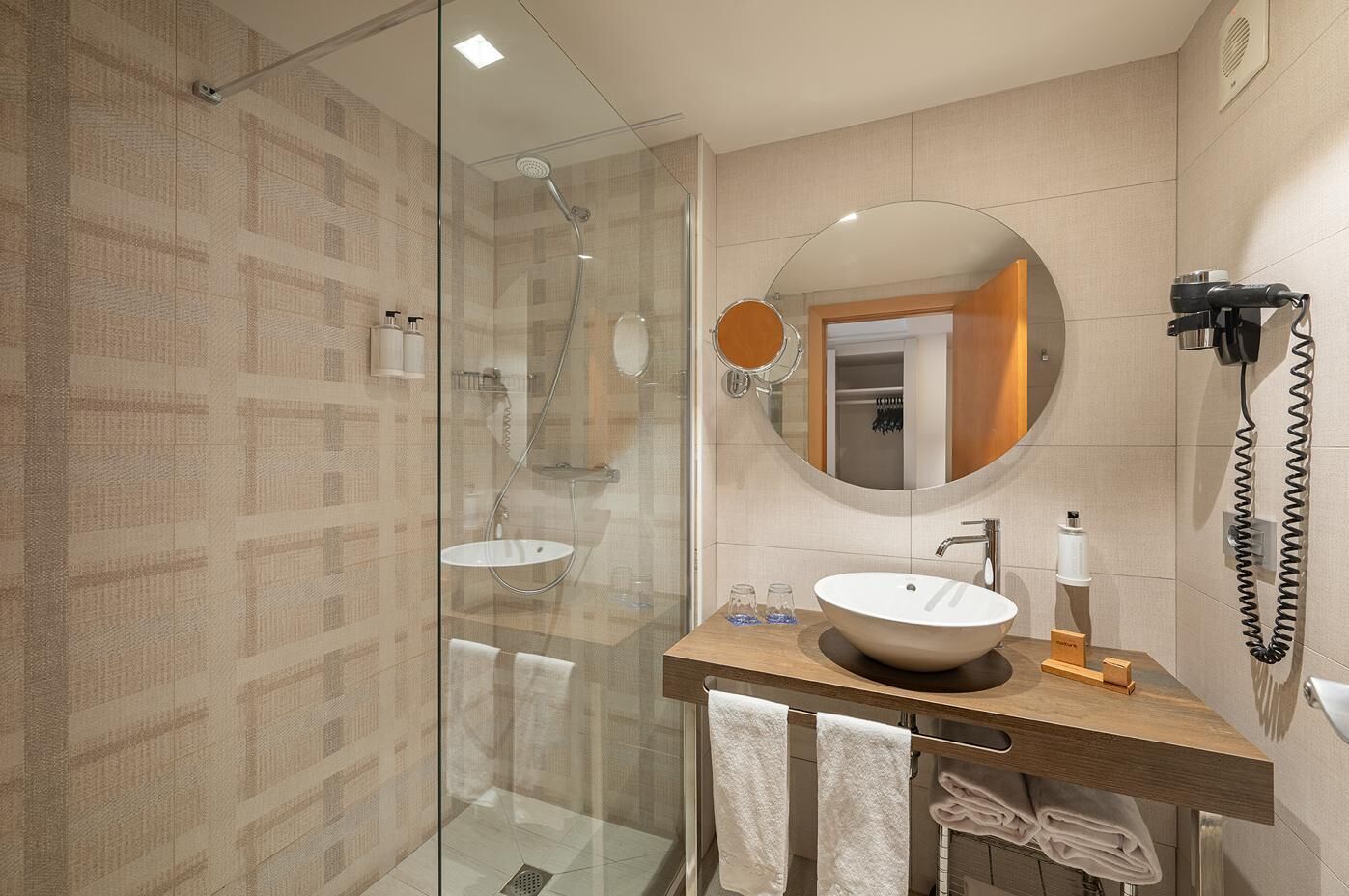 Un baño moderno con ducha de vidrio, tocador de madera con lavamanos grande y espejo redondo.