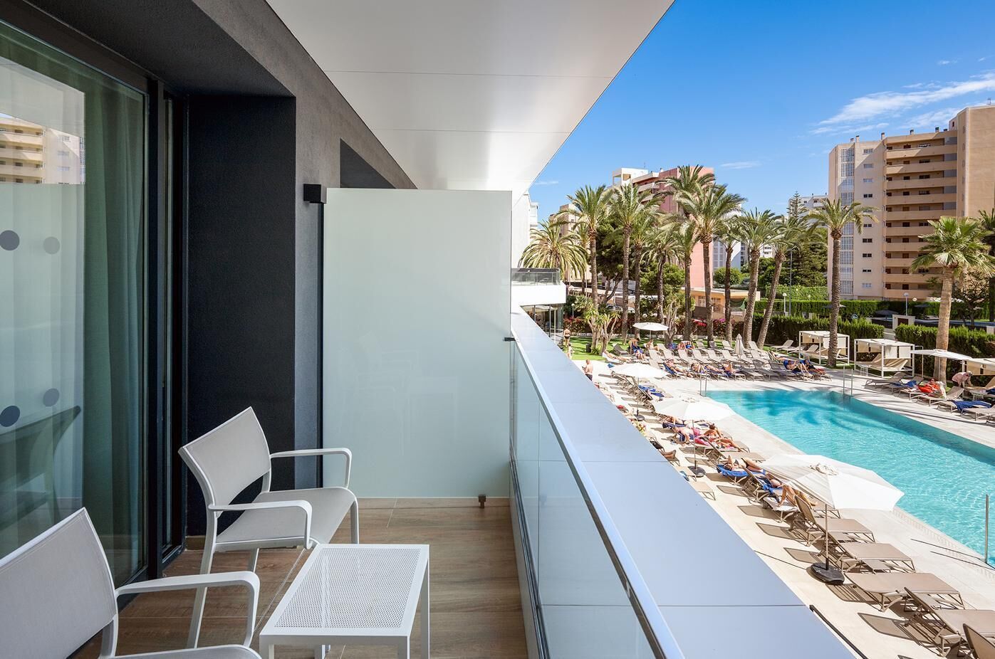 A balcony with white chairs and a table, overlooking a pool with lounge chairs and palm trees.