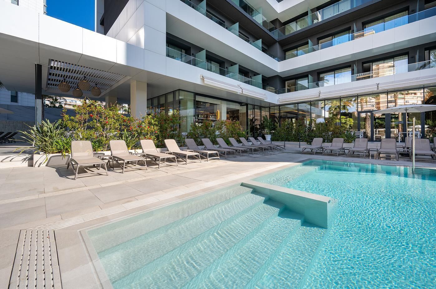 A modern hotel with a large outdoor pool, sun loungers, and a multi-story building with balconies.