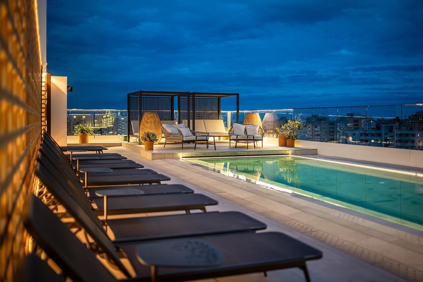 Un área de piscina en la azotea con sillones de descanso, un espacio de asientos con cojines y vista panorámica de la ciudad al anochecer.