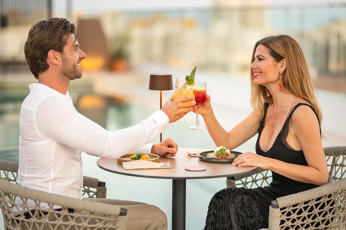 Una pareja cenando en una mesa al aire libre con vistas a la ciudad, sosteniendo bebidas.