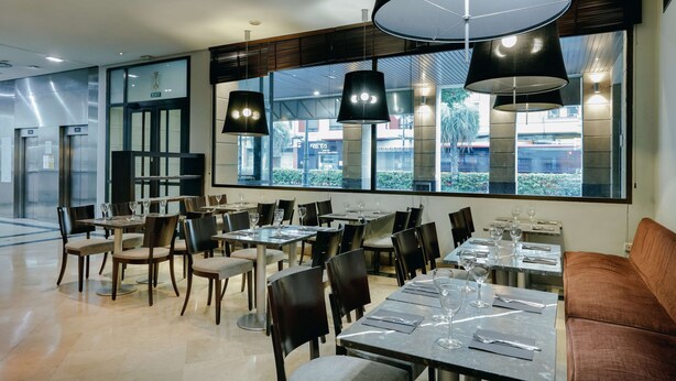 Un restaurante moderno con mesas y sillas, ventanas amplias y lámparas colgantes.