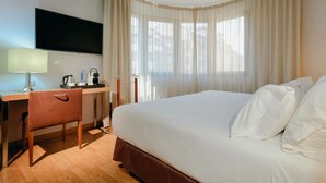 Una habitación de hotel con cama, escritorio con lámpara, televisión y ventana con cortinas.