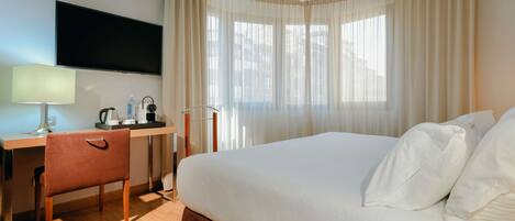 A hotel room with a bed, a desk with a lamp, a television, and a window with curtains.