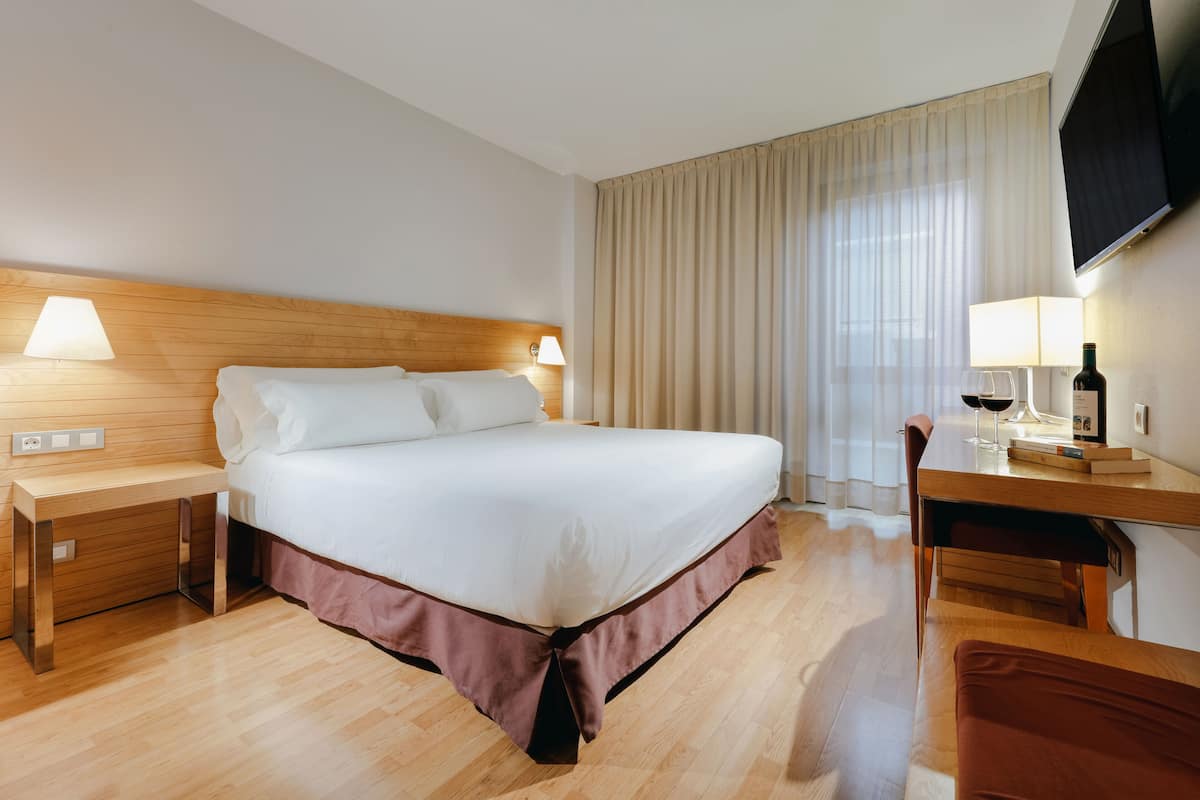 A hotel room with a large bed, wooden headboard, bedside lamps, a desk with a chair, and a TV mounted on the wall.