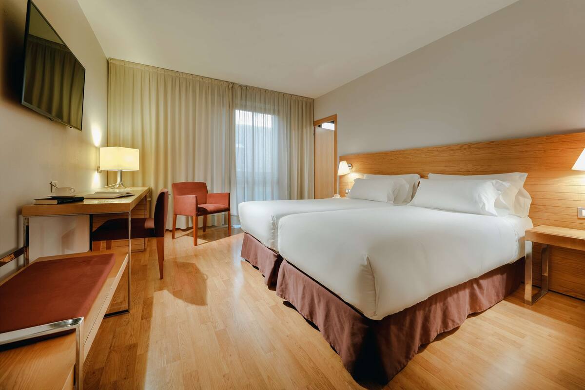 A hotel room with a large bed, a desk with a chair, a TV, and a window with curtains.