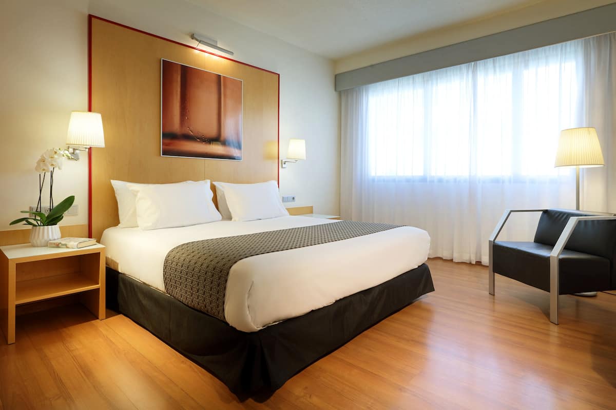 Een hotelkamer met een groot bed, twee bedlampen, een stoel, een kleine tafel met een plant en een schilderij aan de muur.