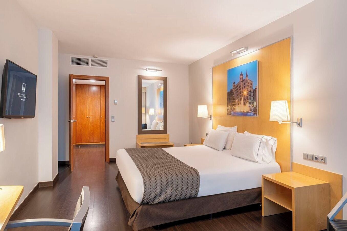 Una habitación de hotel con una cama grande, un cabecero de madera, un televisor de pantalla plana, un espejo y un cuadro de un edificio.