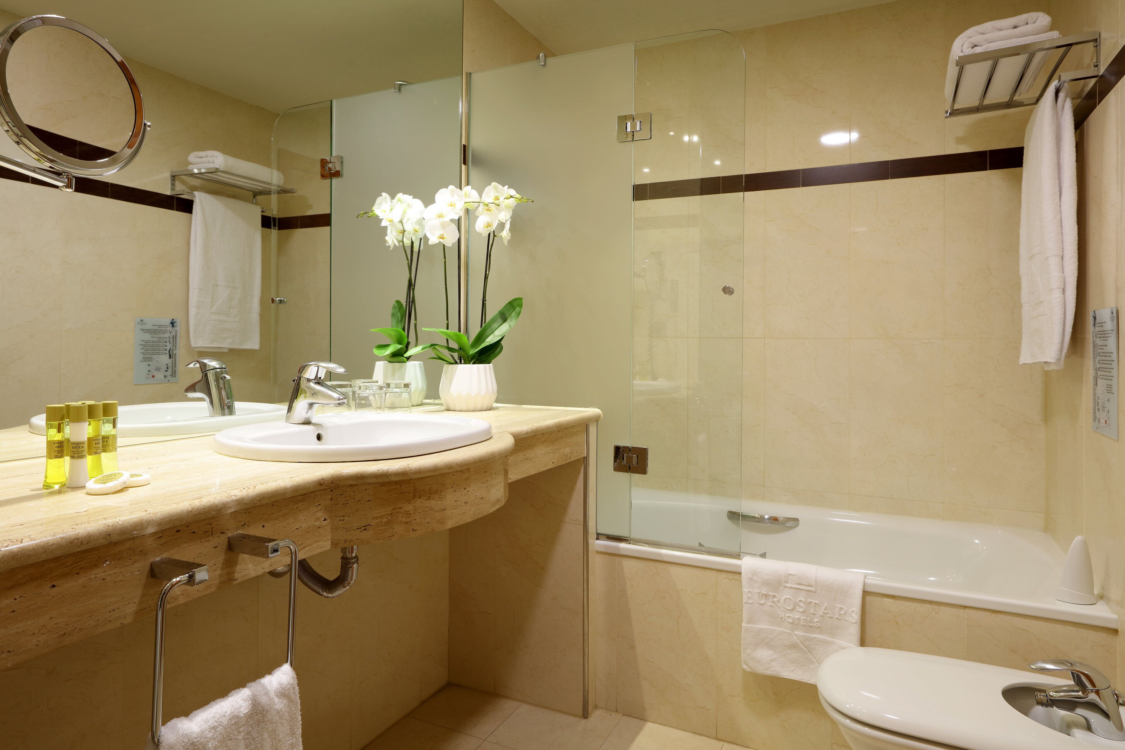 A modern bathroom with a sink, mirror, and bathtub. There are white towels and a vase with white flowers on the counter.