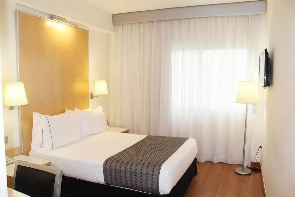 Hotelkamer met een bed, twee bedlampen, een flatscreen televisie, een bureau met stoel en een groot raam met gordijnen.
