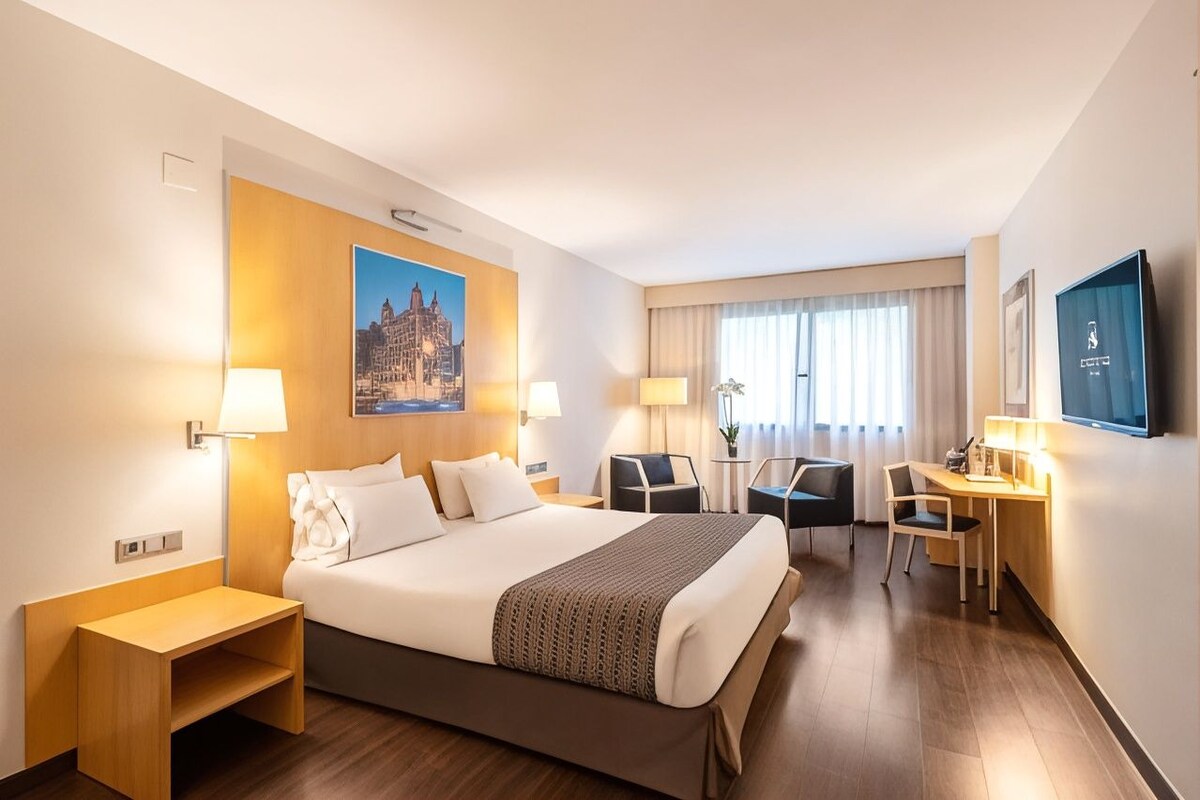 Habitación de hotel con cama, mesitas de noche, escritorio, silla, televisor y ventana con cortinas.