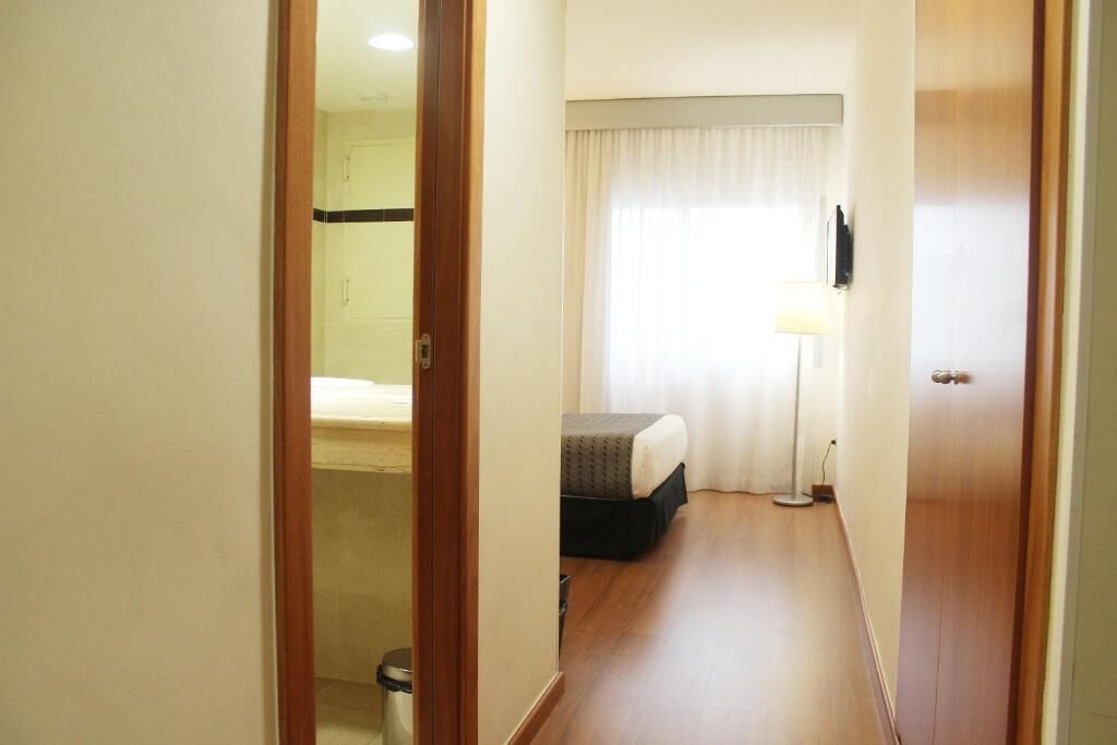A hotel room with a bed, a bedside table, a lamp, and a window with curtains.