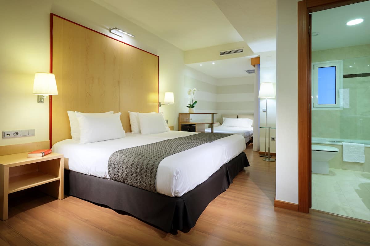 Een hotelkamer met een groot bed, nachtlampjes, een nachtkastje, een kleintje tafeltje met een vaas, en uitzicht op de badkamer.