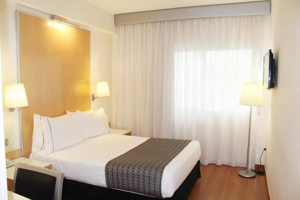 Hotelkamer met een bed, twee bedlampen, een flatscreen televisie, een bureau met lamp en een groot raam met gordijnen.