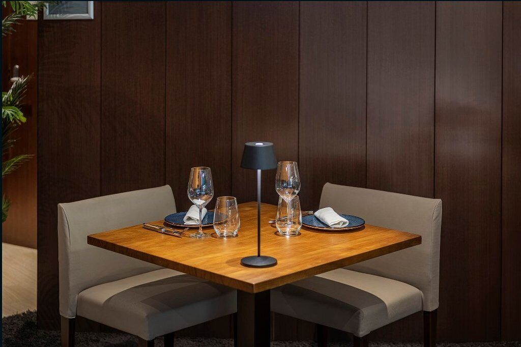 Mesa de jantar posta com quatro taças de vinho, um abajur e pratos, rodeada por cadeiras acolchoadas.