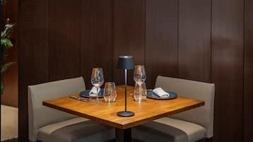 Mesa de jantar posta com quatro taças de vinho, um abajur e pratos, rodeada por cadeiras acolchoadas.