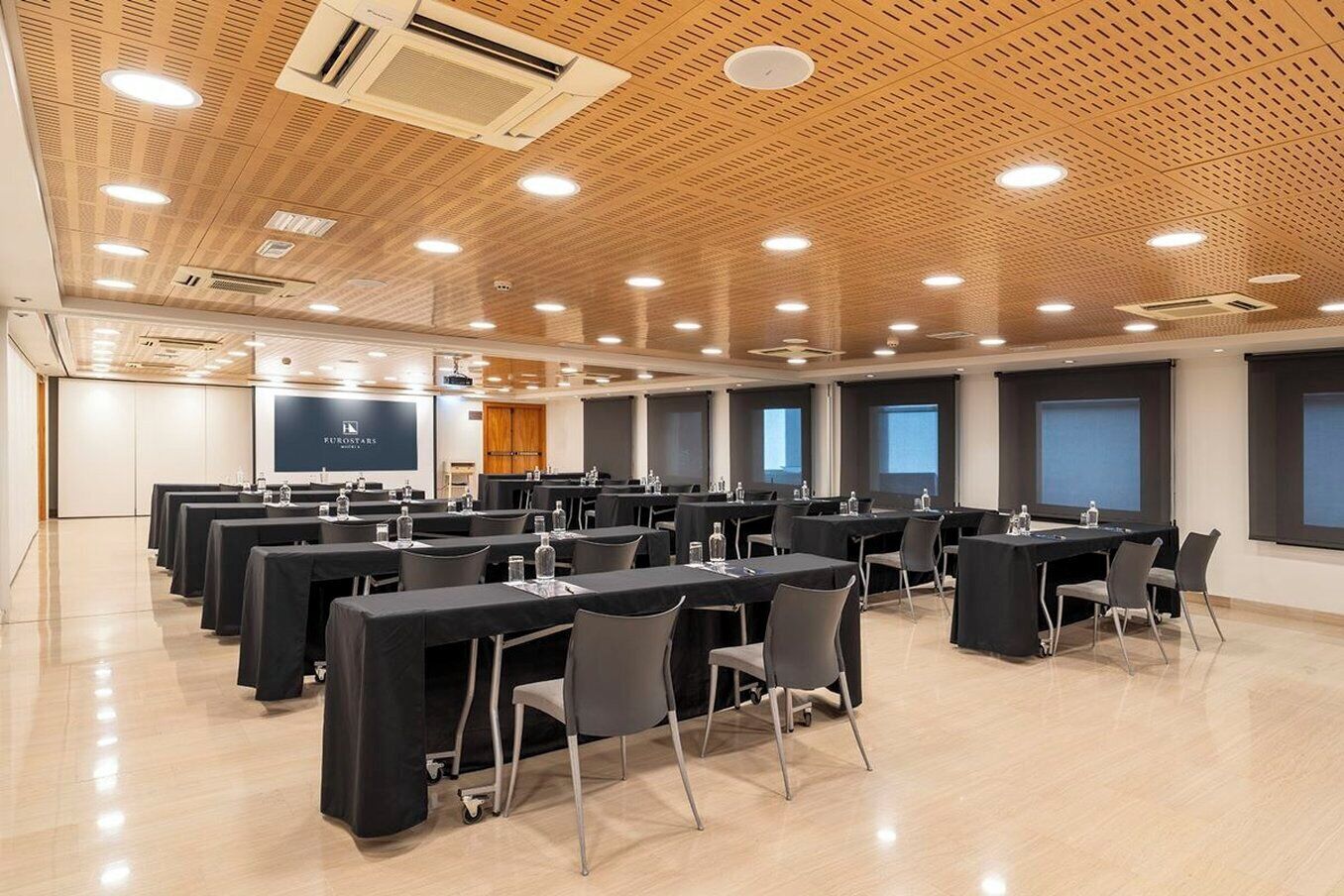 A conference room with multiple tables and chairs, a projector screen, and a wooden ceiling.