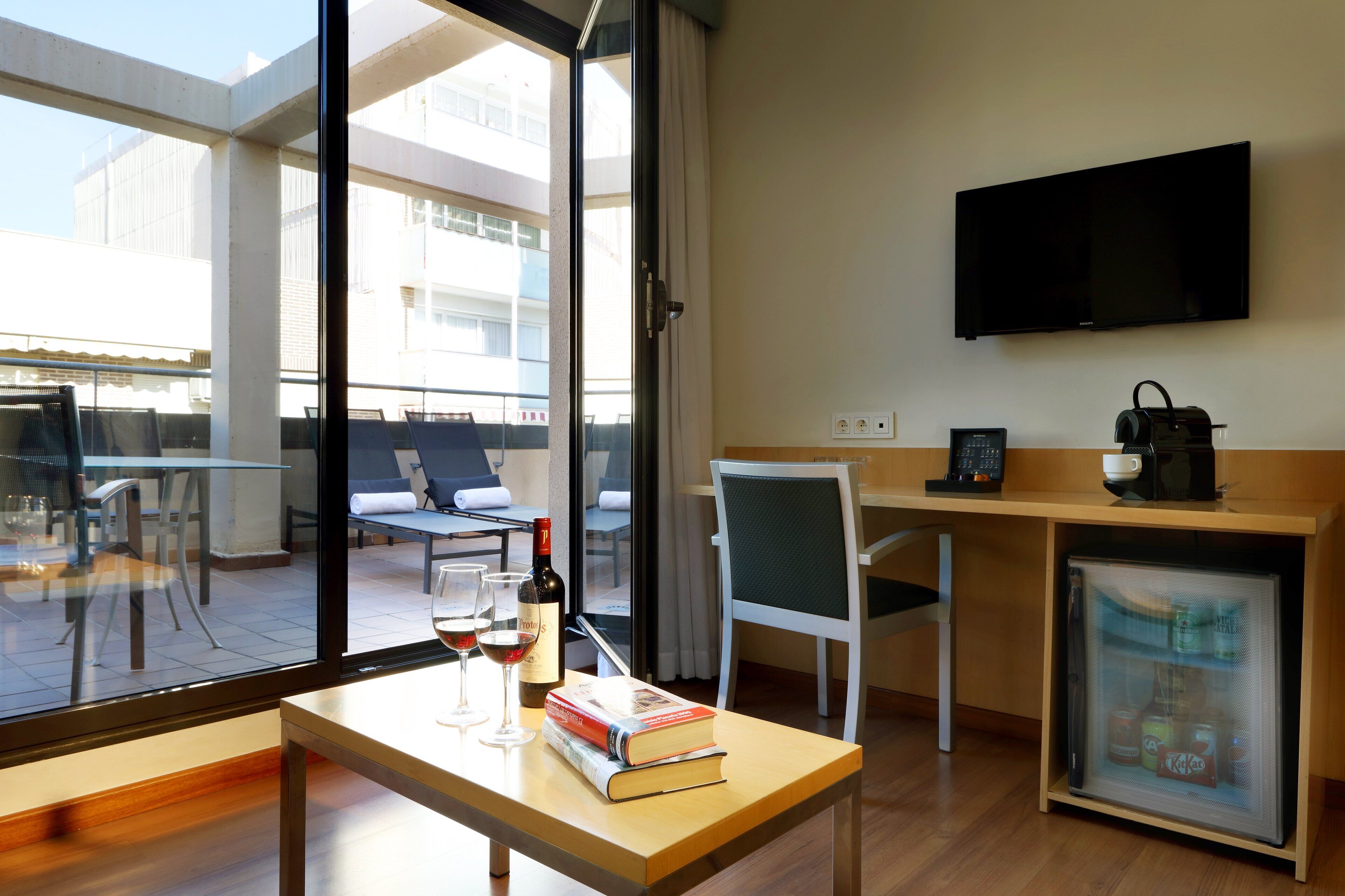 Quarto de hotel moderno com uma janela grande, uma escrivaninha com um computador, uma cadeira, um refrigerador pequeno e uma TV de tela plana.