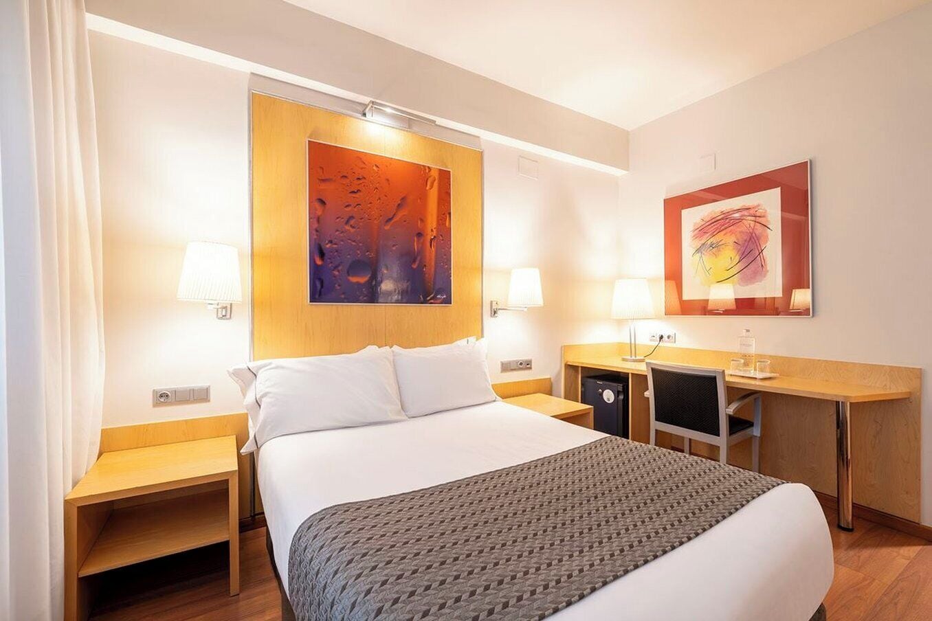Une chambre d’hôtel comprenant un lit, des tables de chevet, un bureau, une chaise, une lampe et des œuvres d’art accrochées aux murs.