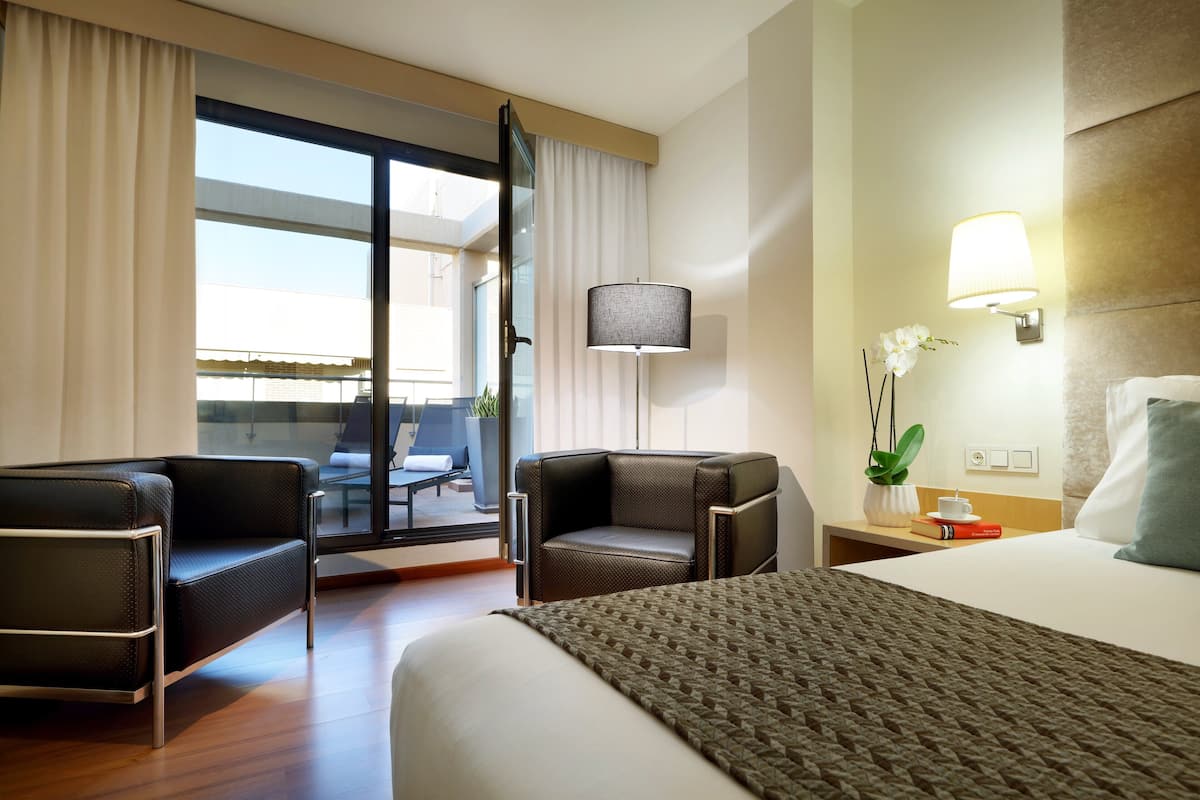 Een hotelkamer met een bed, twee fauteuils, een tafeltje met een lamp, een bloempot en uitzicht op het balkon.