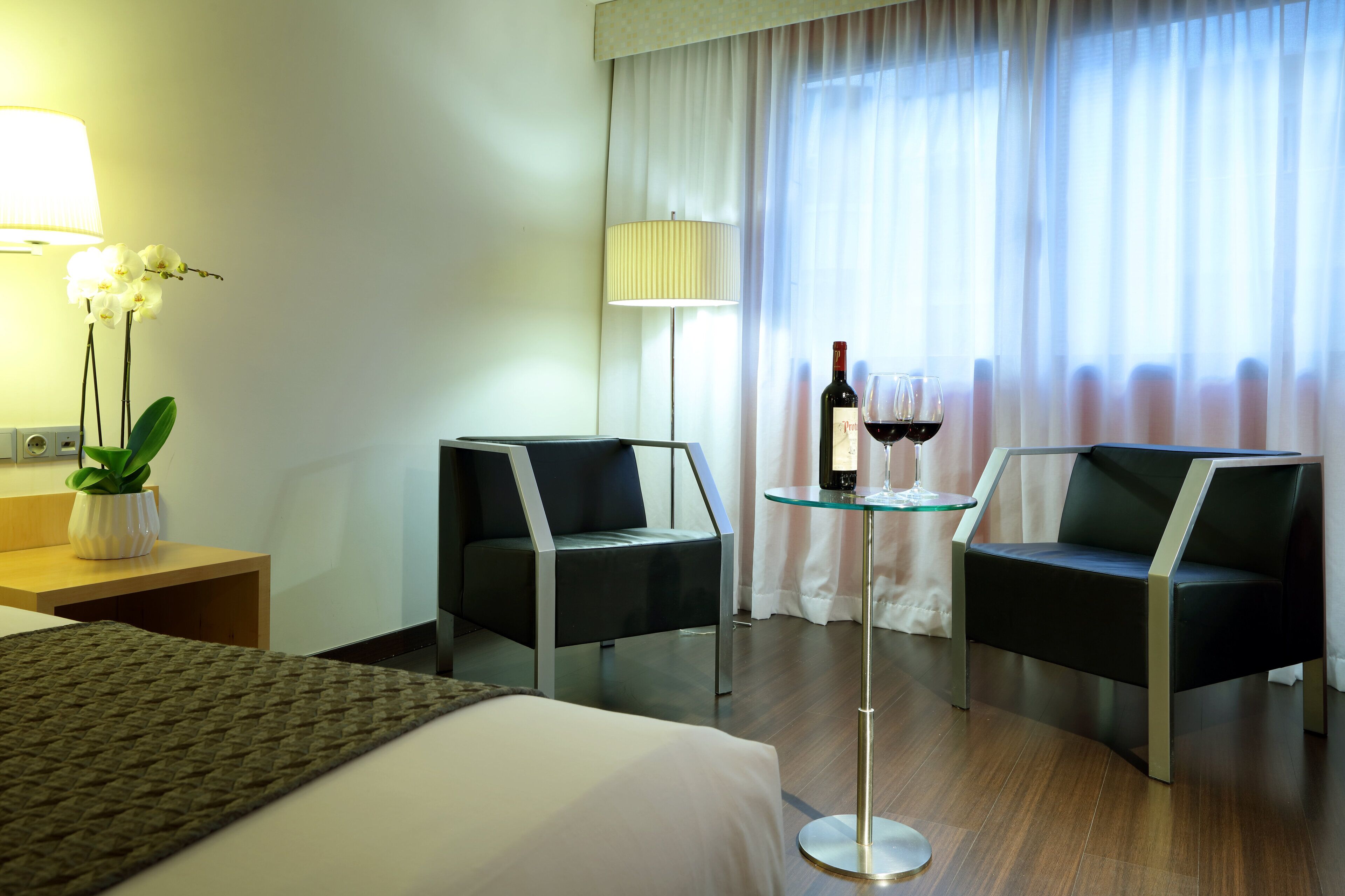 A hotel room with a bed, two armchairs, a small table with a bottle of wine and a glass, a lamp, and a potted plant.