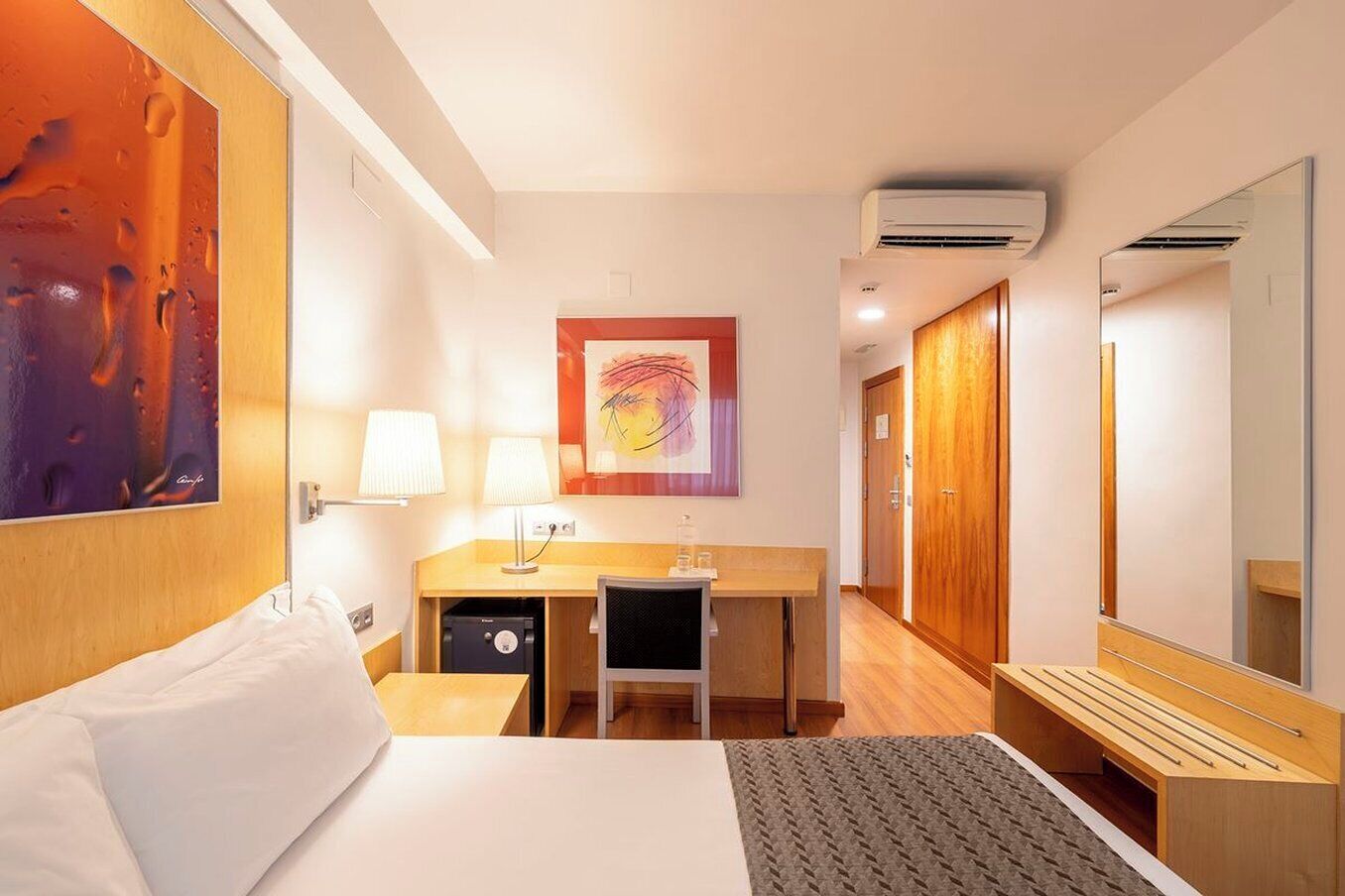A hotel room with a bed, desk, chair, and a large mirror.