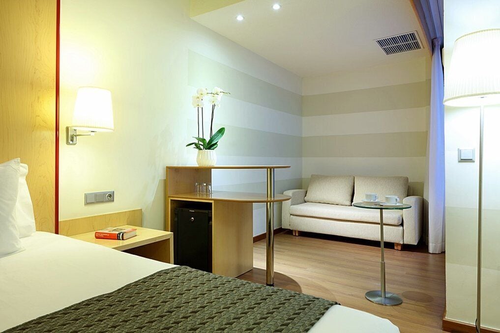 A hotel room with a bed, bedside table, lamp, desk, chair, sofa, and a small potted plant.