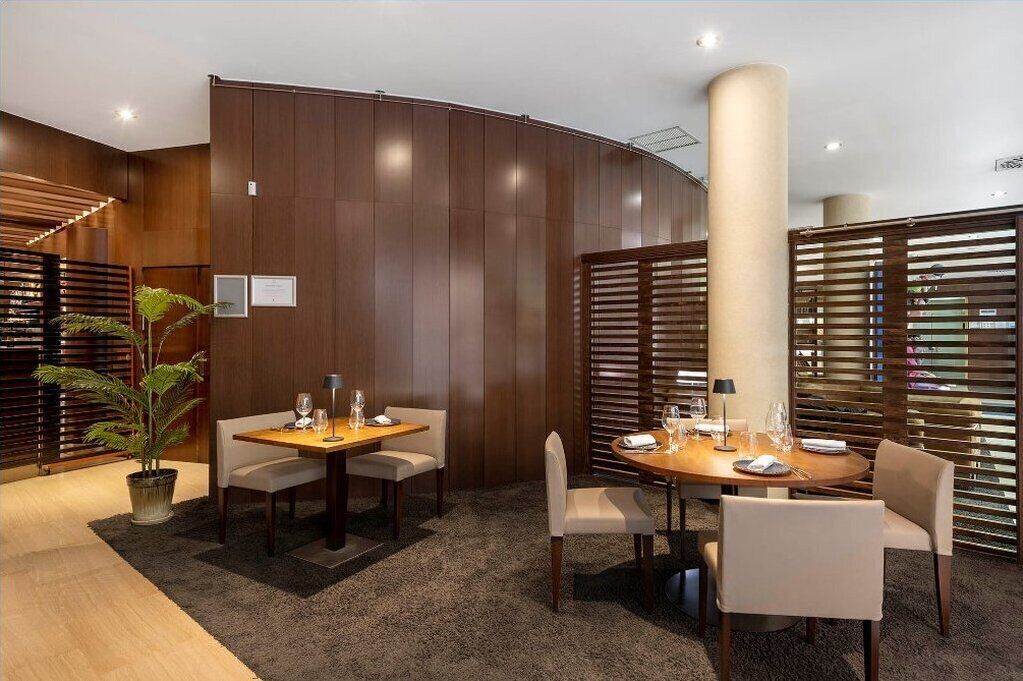 A modern hotel room with a curved wooden wall, a round dining table with chairs, and a potted plant.