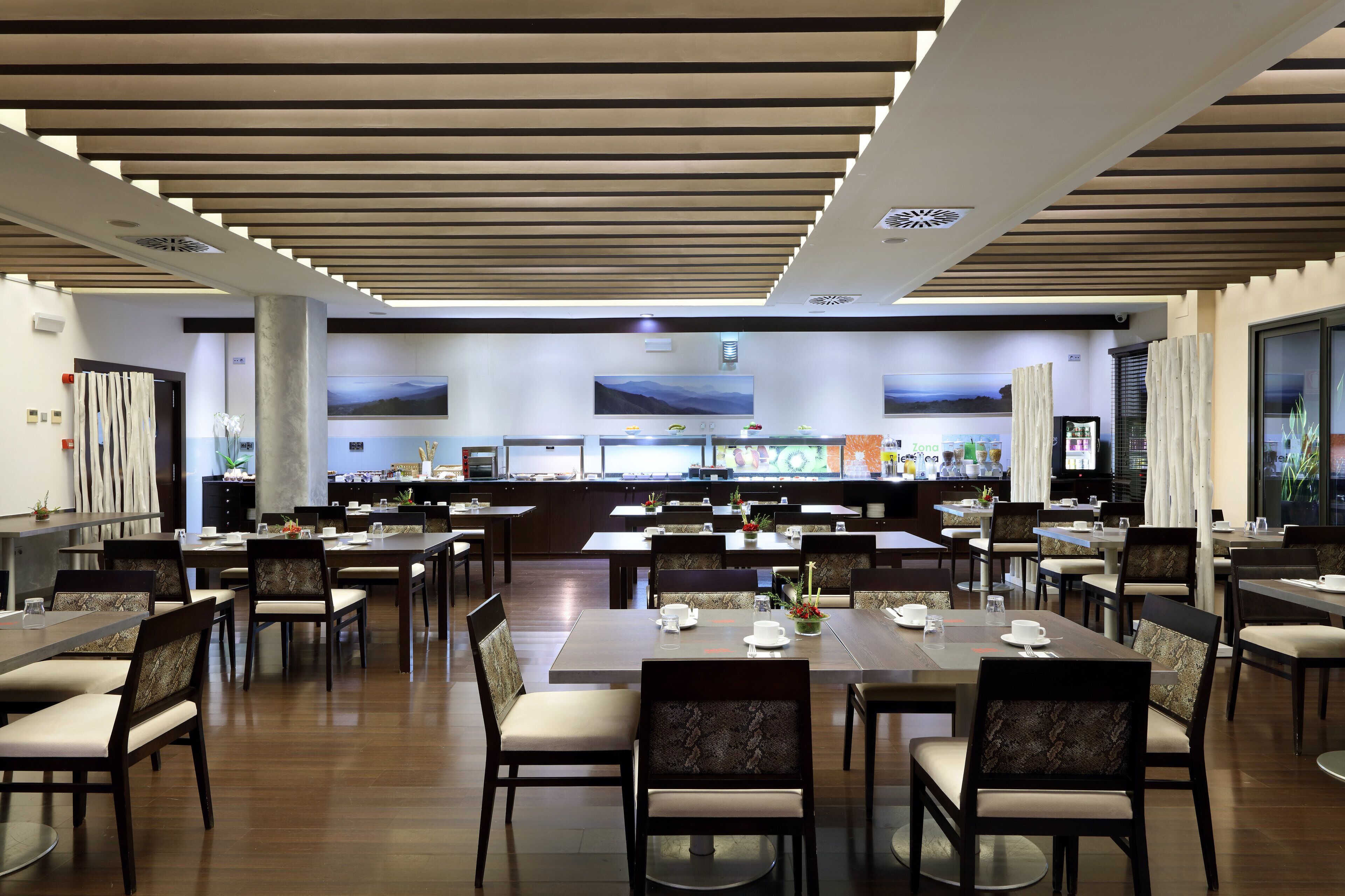 Un vaste espace dédié à la salle à manger, avec plusieurs tables et chaises, un design moderne au niveau du plafond et une vue sur la cuisine en arrière-plan.