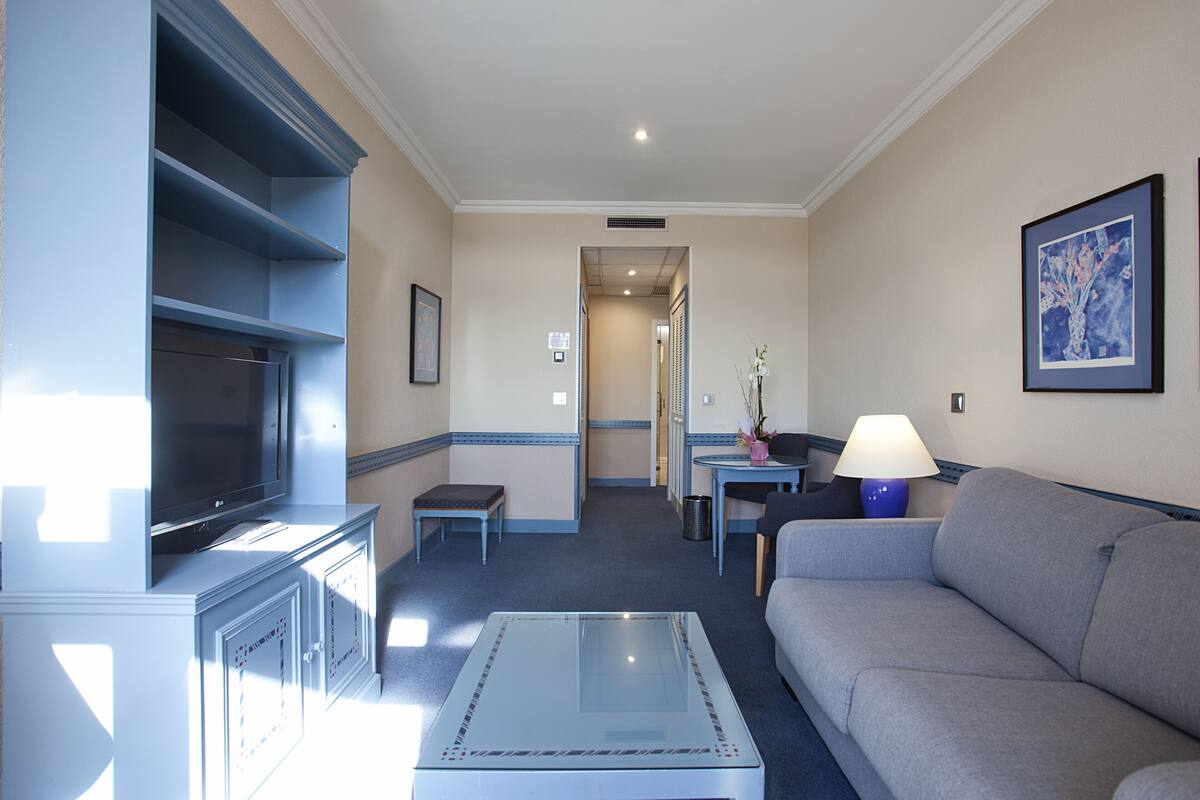 A hotel room with a flat-screen TV, a sofa, a glass coffee table, a small table with a lamp, and a framed picture on the wall.