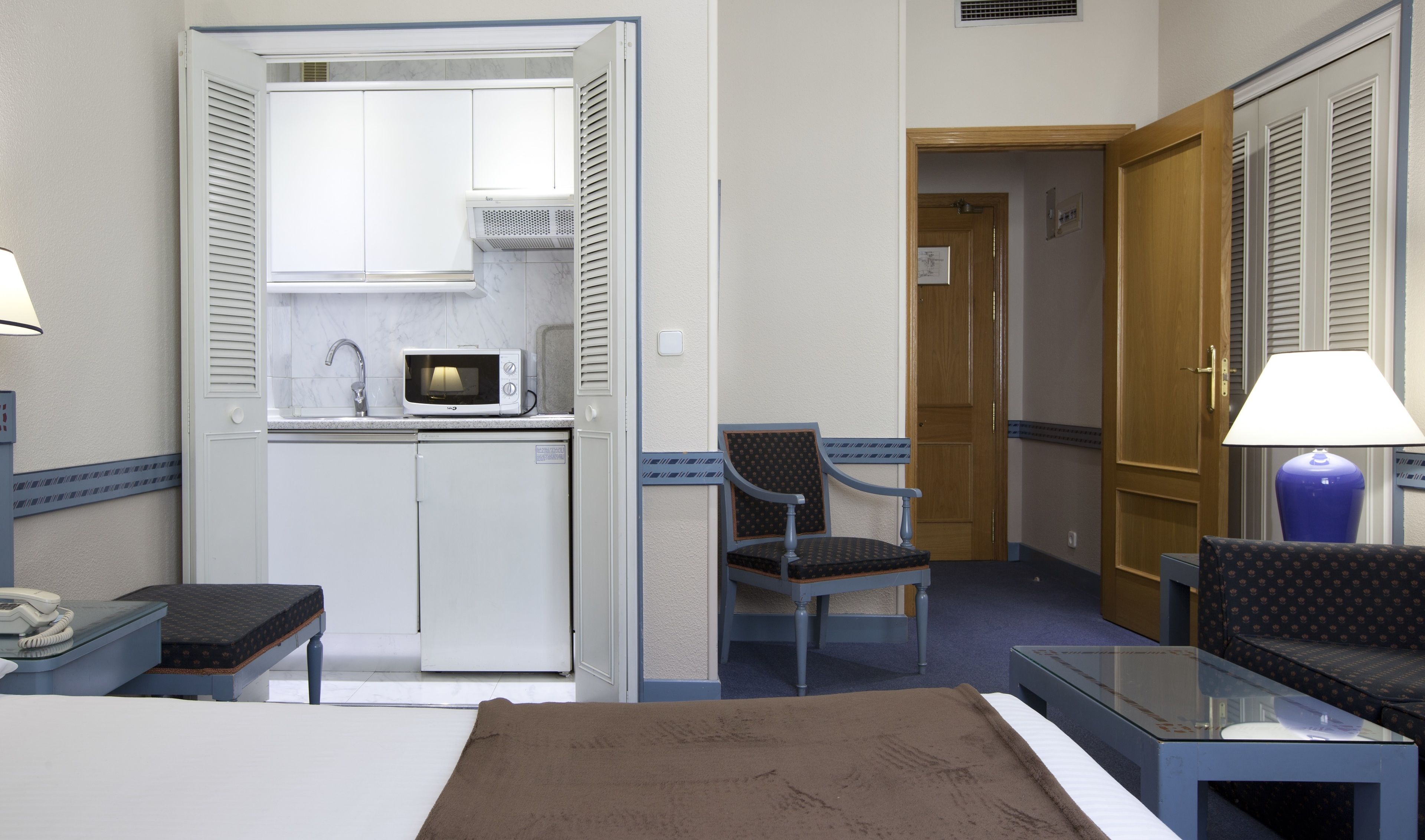 A compact hotel room with a kitchenette, a bed, a small table, a chair, and a lamp.