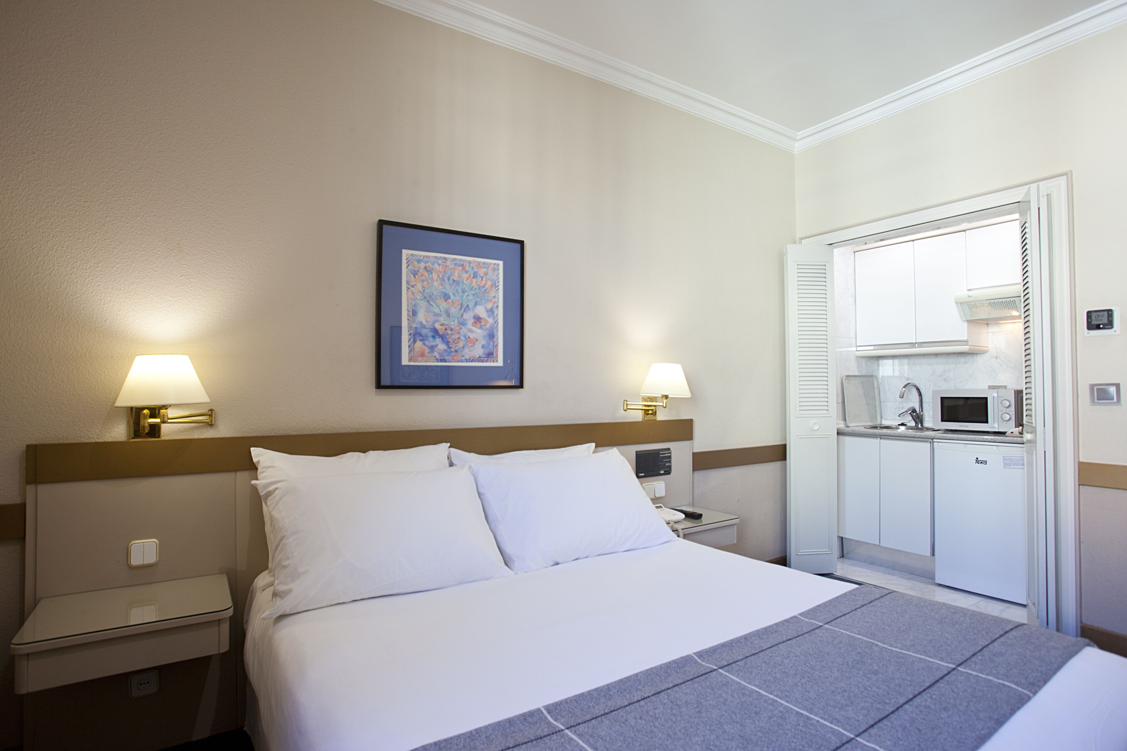 Una habitación de hotel con cama, lámparas de noche, un cuadro, microondas y kitchenette.