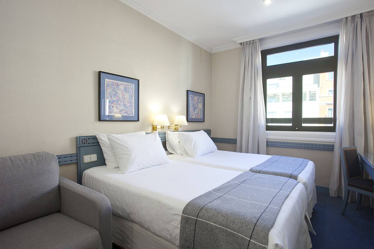 A hotel room with two beds, a desk, and a chair. There are two framed pictures on the wall and a window with curtains.