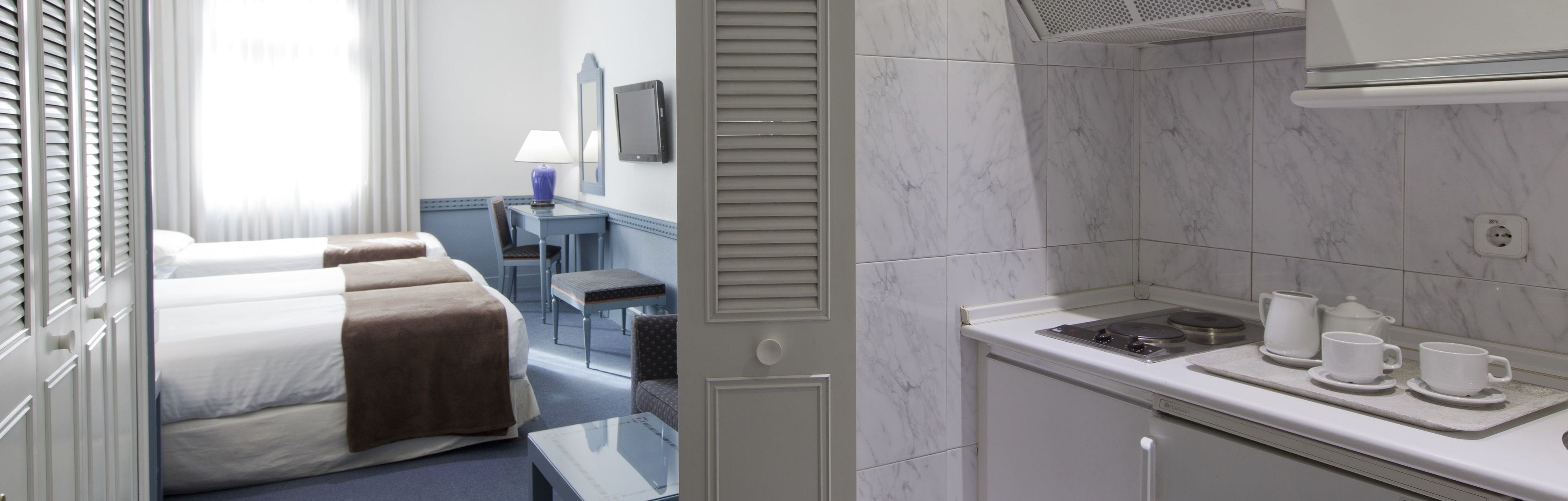 Una habitación de hotel con cama, mesita de noche y una pequeña kitchenette equipada con estufa y tazas.