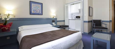 A hotel room with a bed, bedside table, lamp, framed artwork, and a chair.