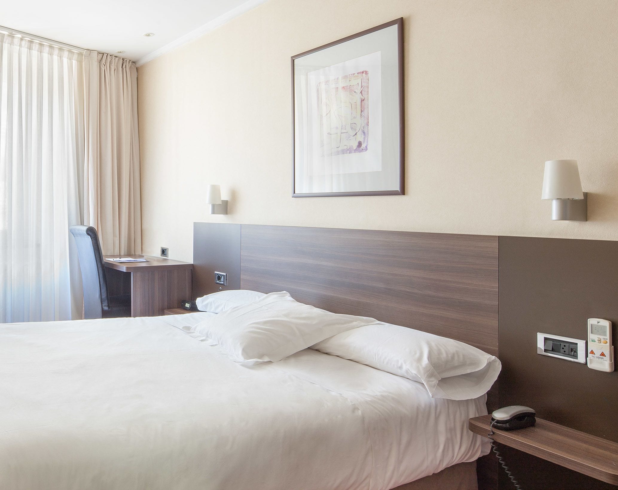 A hotel room with a large bed, a desk with a chair, a bedside table, a wall-mounted lamp, and a framed picture on the wall.