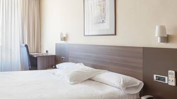 A hotel room with a large bed, a desk with a chair, a bedside table, a wall-mounted lamp, and a framed picture on the wall.