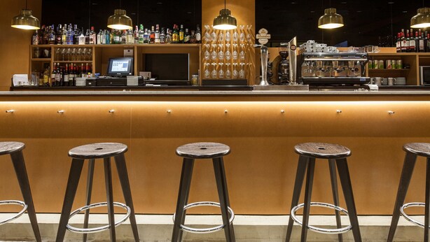 Zona de bar con taburetes, una barra con un ordenador y estantes llenos de botellas de licor.